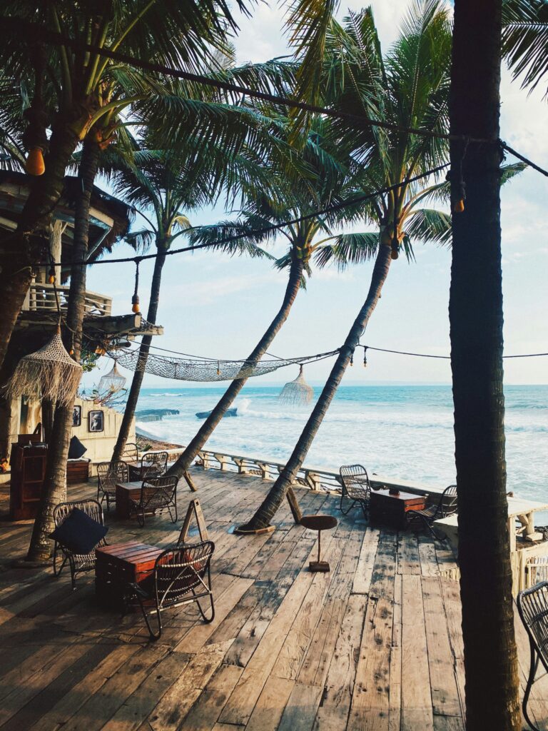 A cafe by the sea with tables and chairs and palm trees
