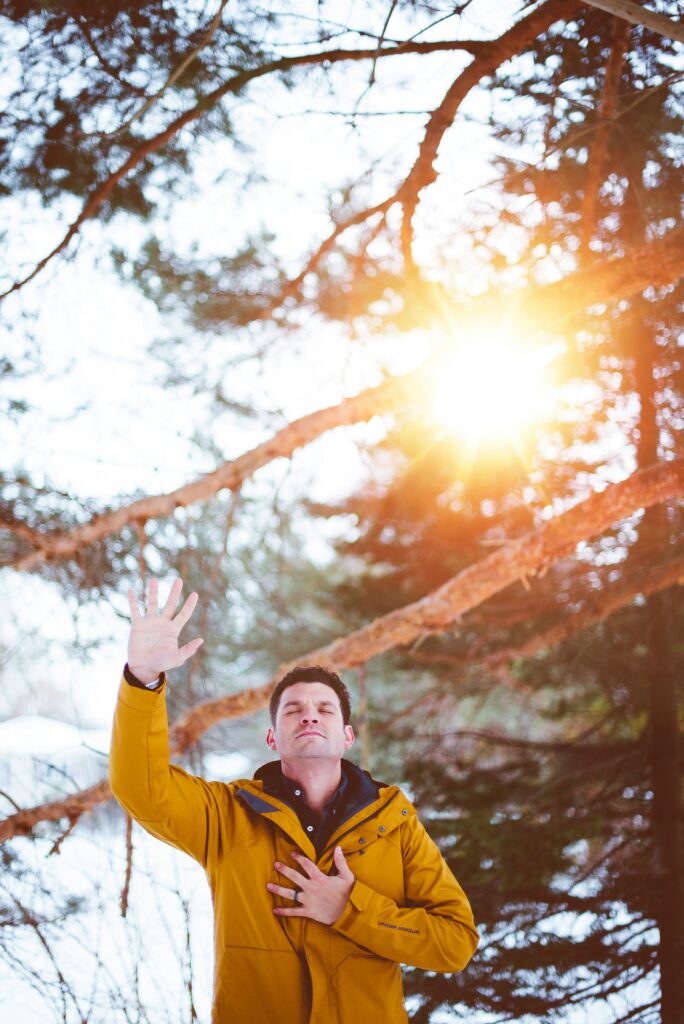 A man stands in the forest with one hand outstretched and his eyes closed in a winter jacket