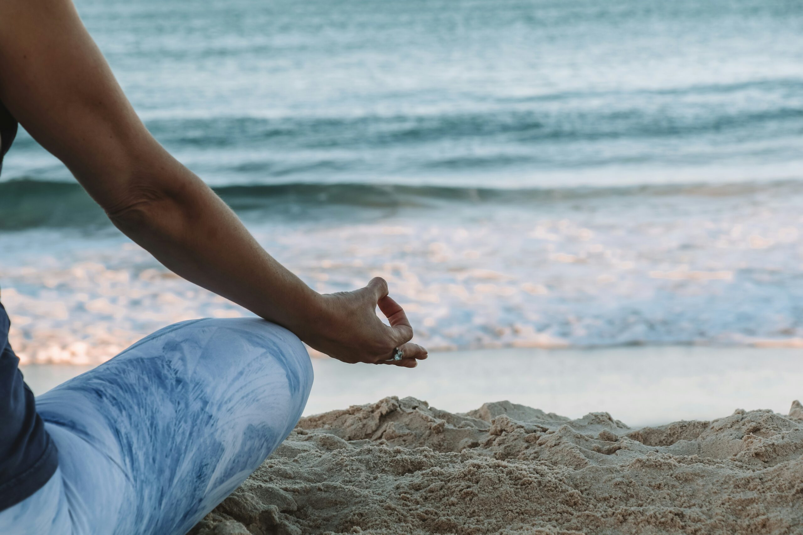 A person meditating on the beach, sitting in a cross-legged position with their hand in a Gyan Mudra gesture