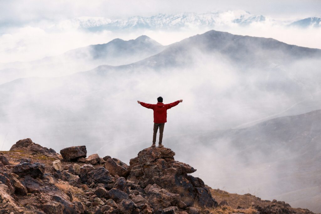 A man stands on the mountain peak with outstretched arms in front of a magnificent natural landscape